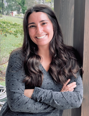 Headshot of Dani Rimbach-Jones. She has light skin and dark long hair. She is weaing a grey sweater and is leaning against a wooden beam. She is smiling in the photo.